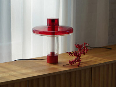 Nevo table lamp with red disc-shaped top and base, clear cylindrical body, and warm modern lighting on a wooden surface